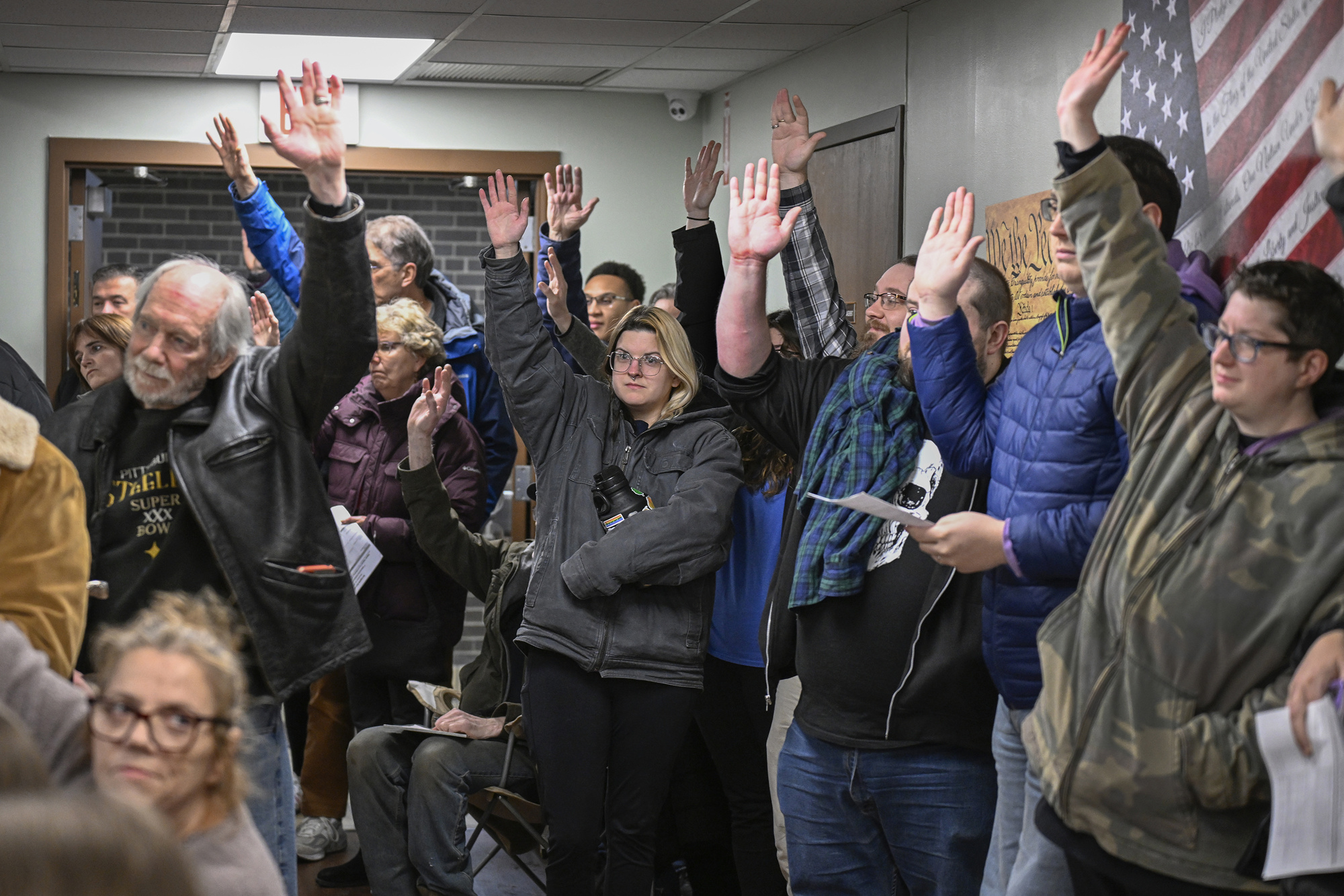 A group of people indoors raise their hands during a meeting or event; most are standing and holding papers, and an American flag is visible on the wall in the background.