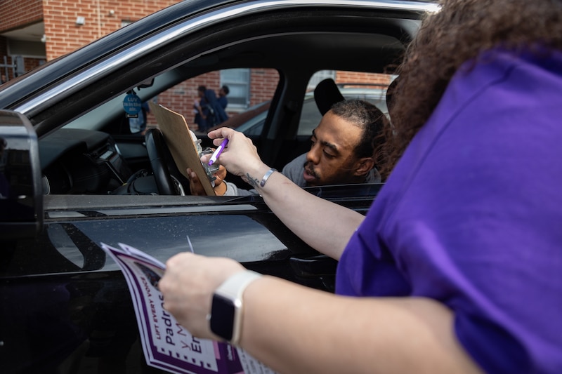 A photograph of a Black man sitting in a car and a woman handing him a clipboard with school info.