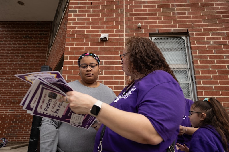 A photograph of two adult women looking at a flyer outside of a brick school building.