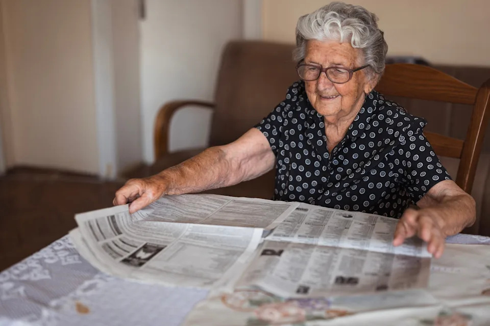 Elderly person with glasses reading a newspaper at a table, smiling contentedly