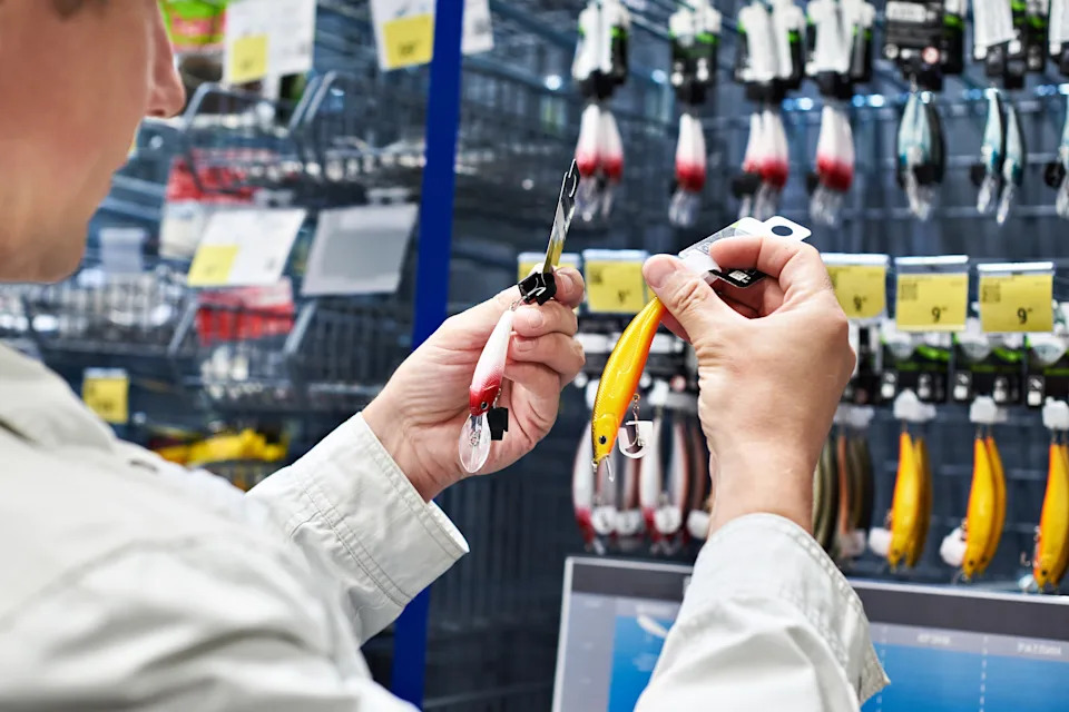Person examines fishing lures in a store aisle, holding a yellow lure in one hand and a multicolored one in the other, with more lures in the background