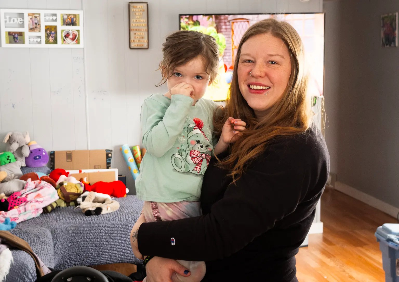 A white woman smiles while holding her daughter, who looks at the camera through a bunched hand.