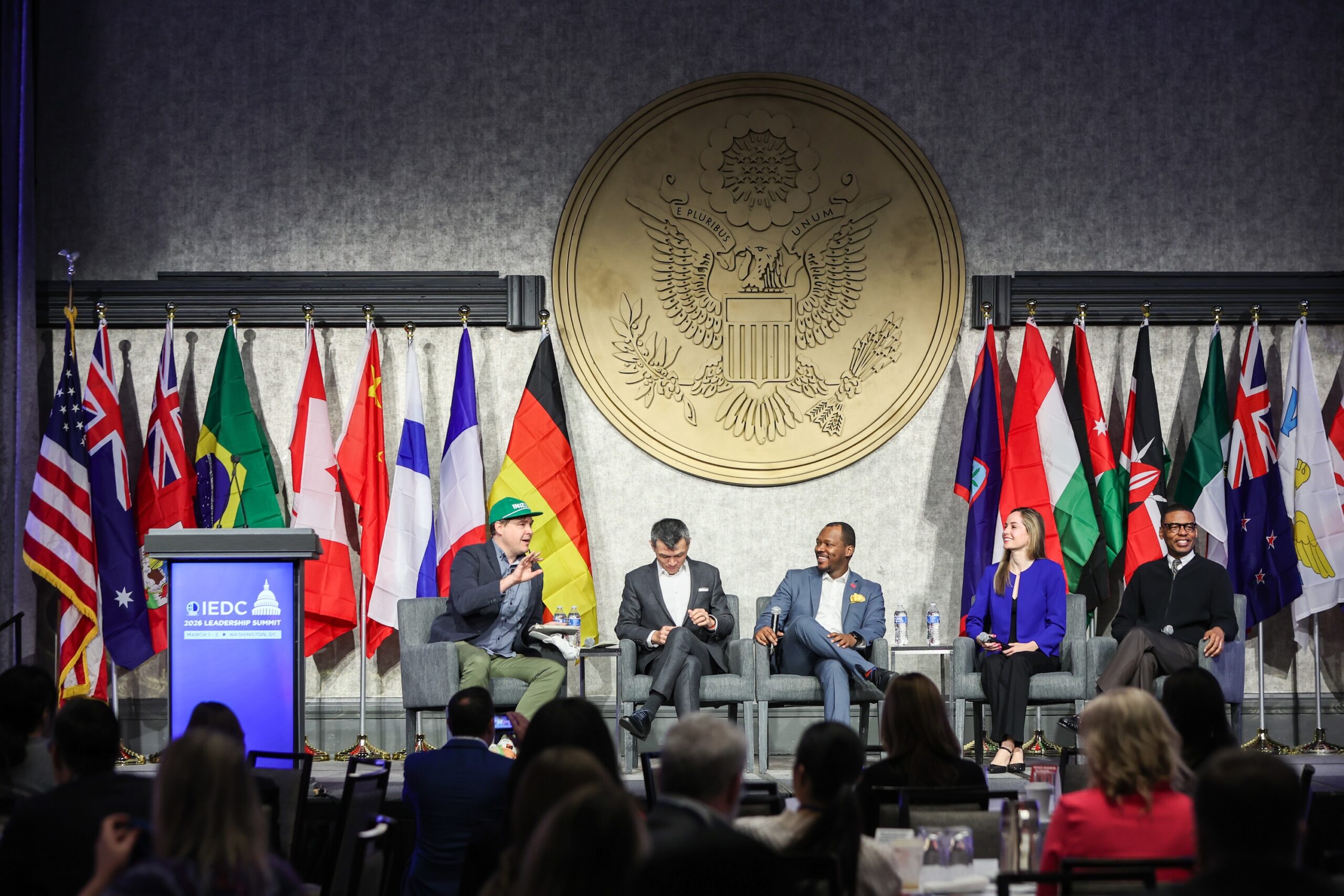 Five people sit on a panel discussion stage with international flags in the background and an audience in the foreground. One panelist wears a green hat.
