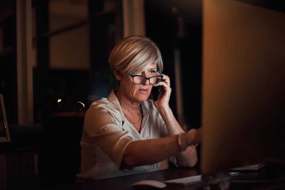 An older adult works at a computer at night, wearing glasses and holding a phone to their ear, focused on the screen