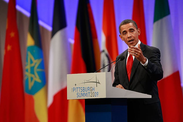 A thin Black man in a suit and red tie speaks at a podium with world flags behind him.