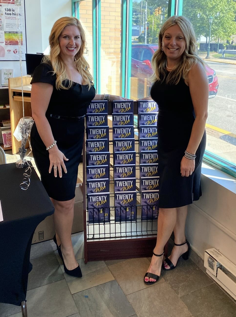 Two women in black dresses stand beside a display of "TWENTY WON" books in front of a window inside a brightly lit room.