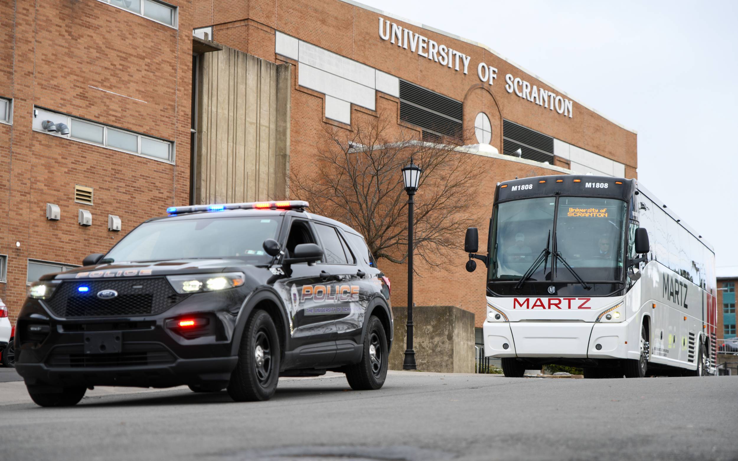 University of Scranton police SUV with flashing lights parked near a Martz coach bus outside a campus building.