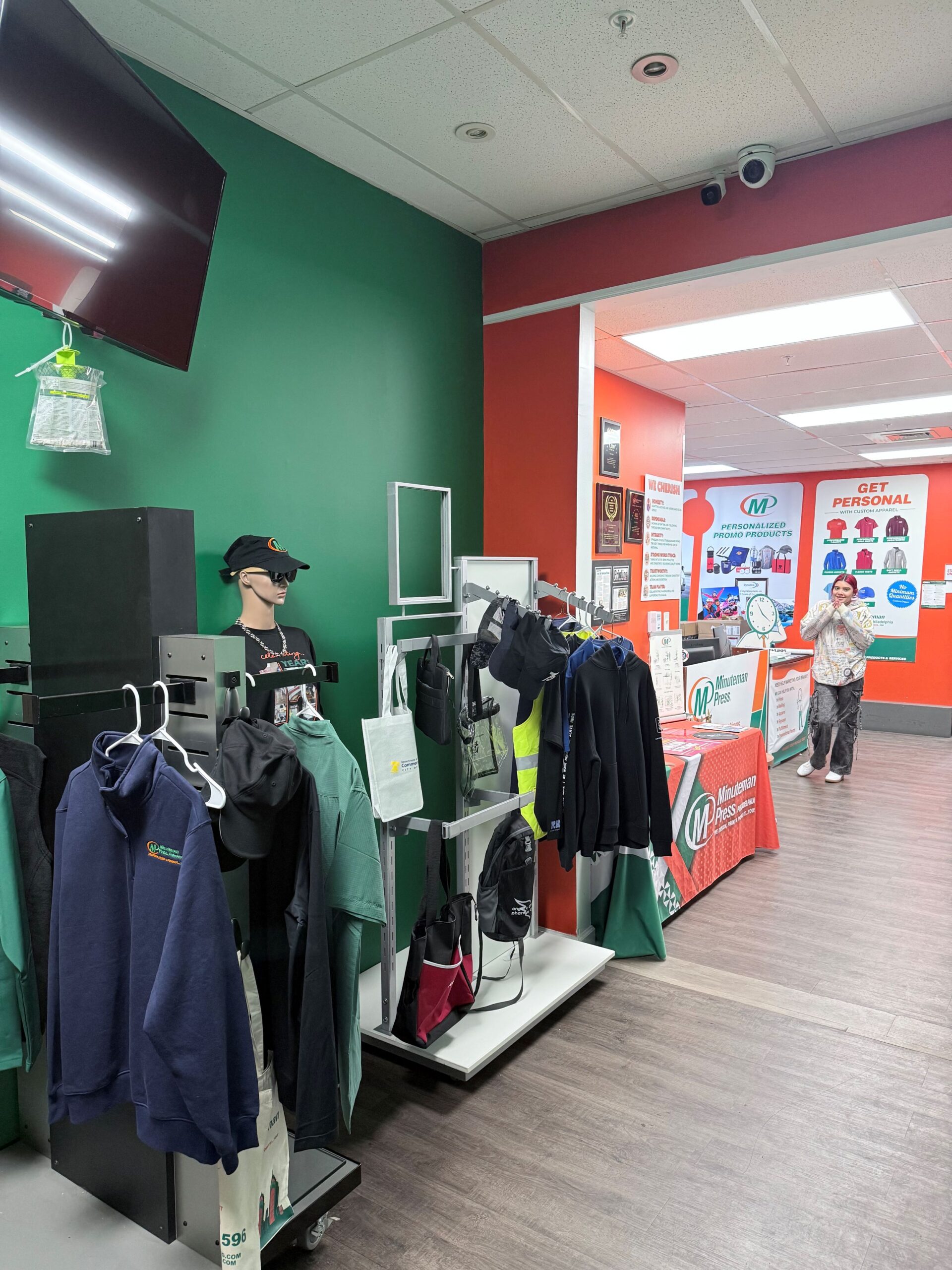 A retail store interior with green and red walls, clothing and accessories on display racks, and a person near a service counter in the background.