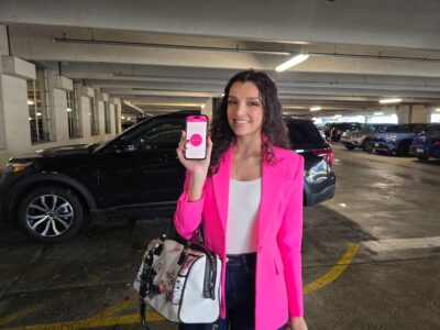Woman in a bright pink blazer stands in a parking garage, smiling and holding up a smartphone displaying an app with a pink interface.