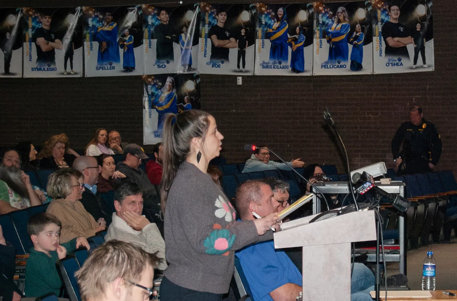 A woman with brown hair stands at a lectern in an auditorium. 