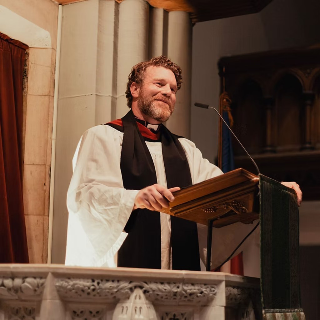 Rev. Aidan Smith, head priest of the Episcopal Church in Pittsburgh, smiling at a lectern.