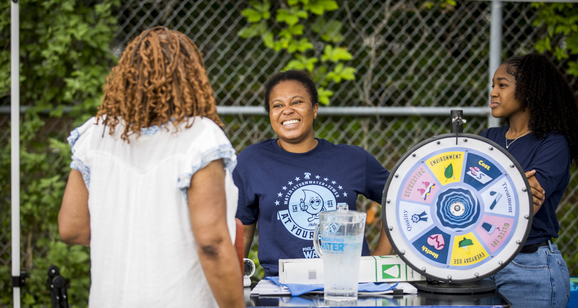Two smiling PWD employees in dark blue PWD t-shirts stand behind the Philly Water Bar table, talking to a patron with shoulder length curly brown hair and a light blue blouse facing away from the camera. A frosty pitcher of Philly tap water and a "wheel of water" sit on top of the table.