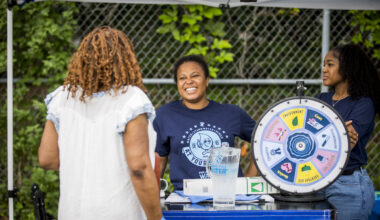 Two smiling PWD employees in dark blue PWD t-shirts stand behind the Philly Water Bar table, talking to a patron with shoulder length curly brown hair and a light blue blouse facing away from the camera. A frosty pitcher of Philly tap water and a "wheel of water" sit on top of the table.
