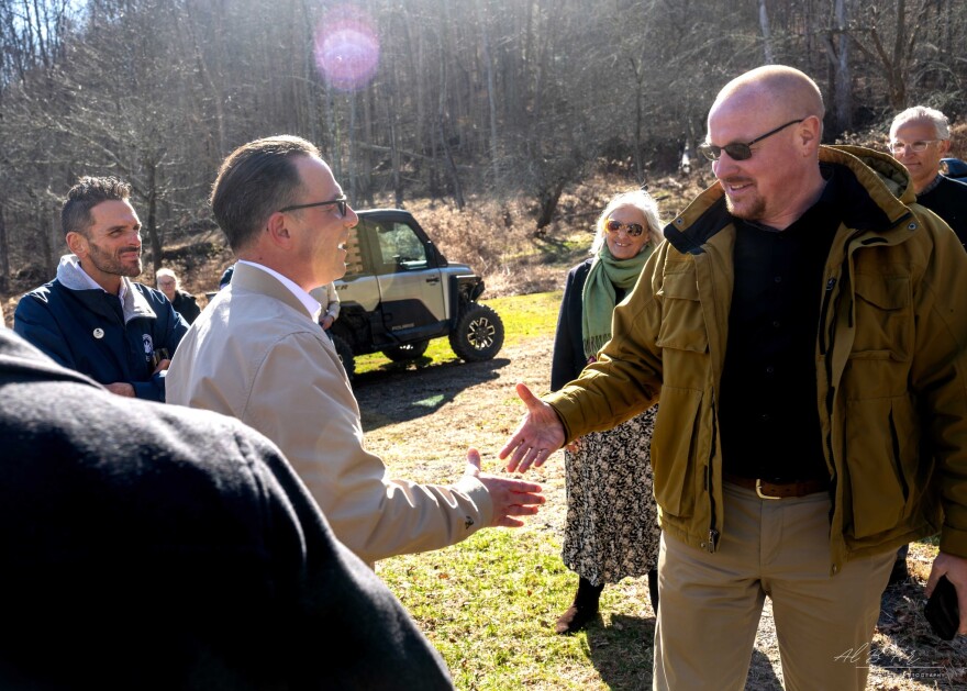 Two men shake hands outside, surrounded by other people in the background.