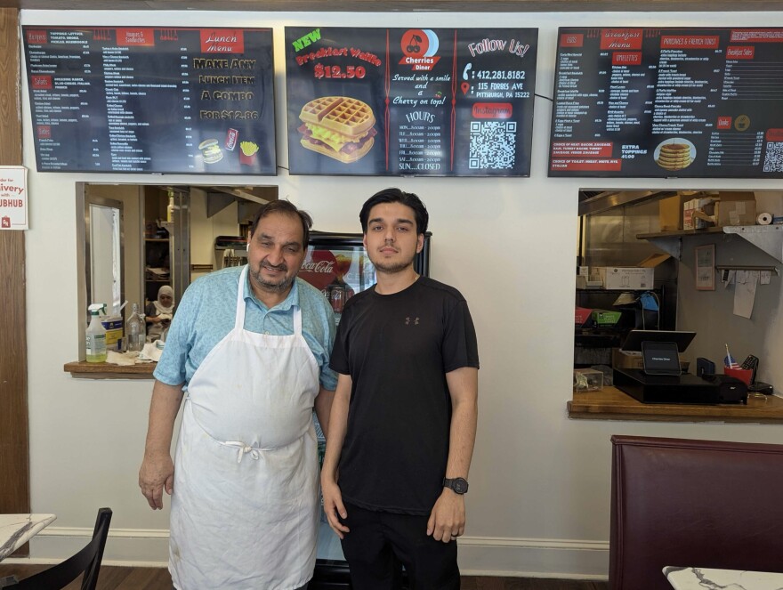 A man wearing an apron and his son stand under a wall-mounted diner menu.