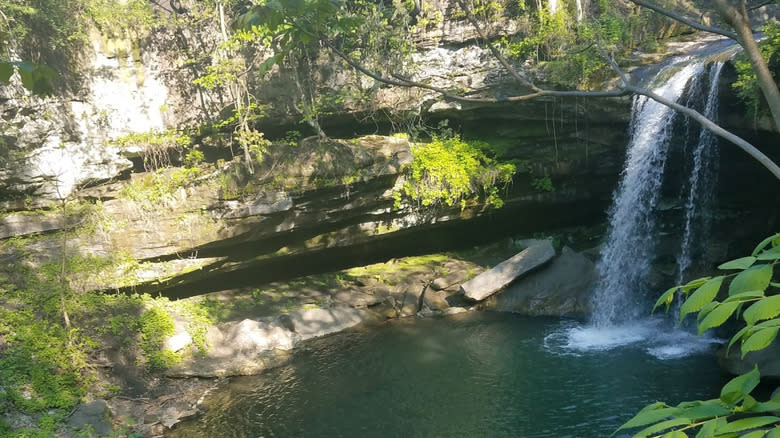 Natural rock walls surrounding a blue-green pool with a waterfall flowing down the right side of the frame