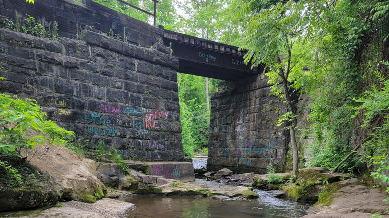 Old railroad bridge over a small stream, with stone walls scrawled in colorful graffiti, surrounded by trees