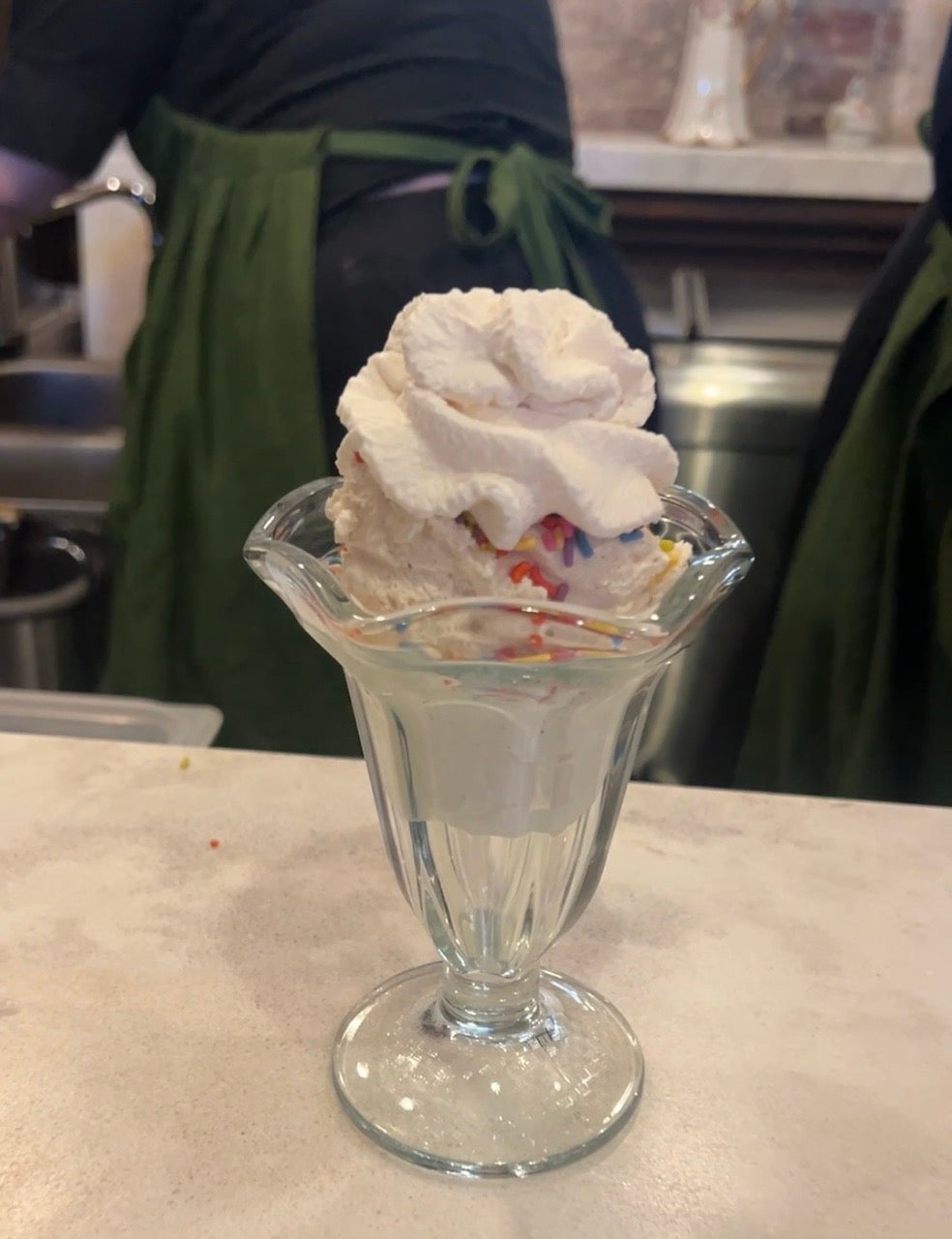 A vanilla ice cream sundae with whipped cream on top in a glass cup sits on a marble counter.