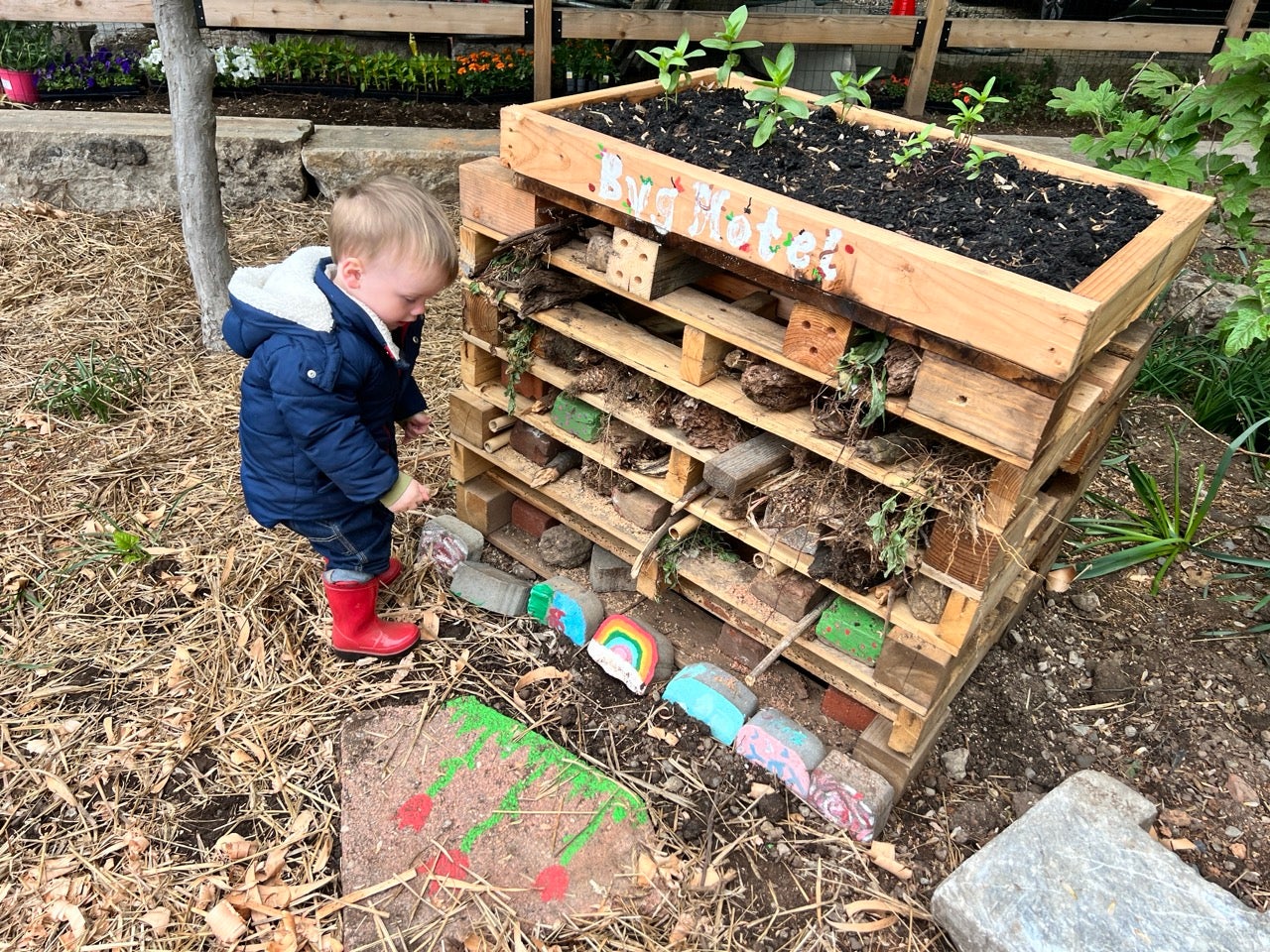 A little boy in red rain boots plays in front of a "Bug Hotel" made of soil and wood pallets.