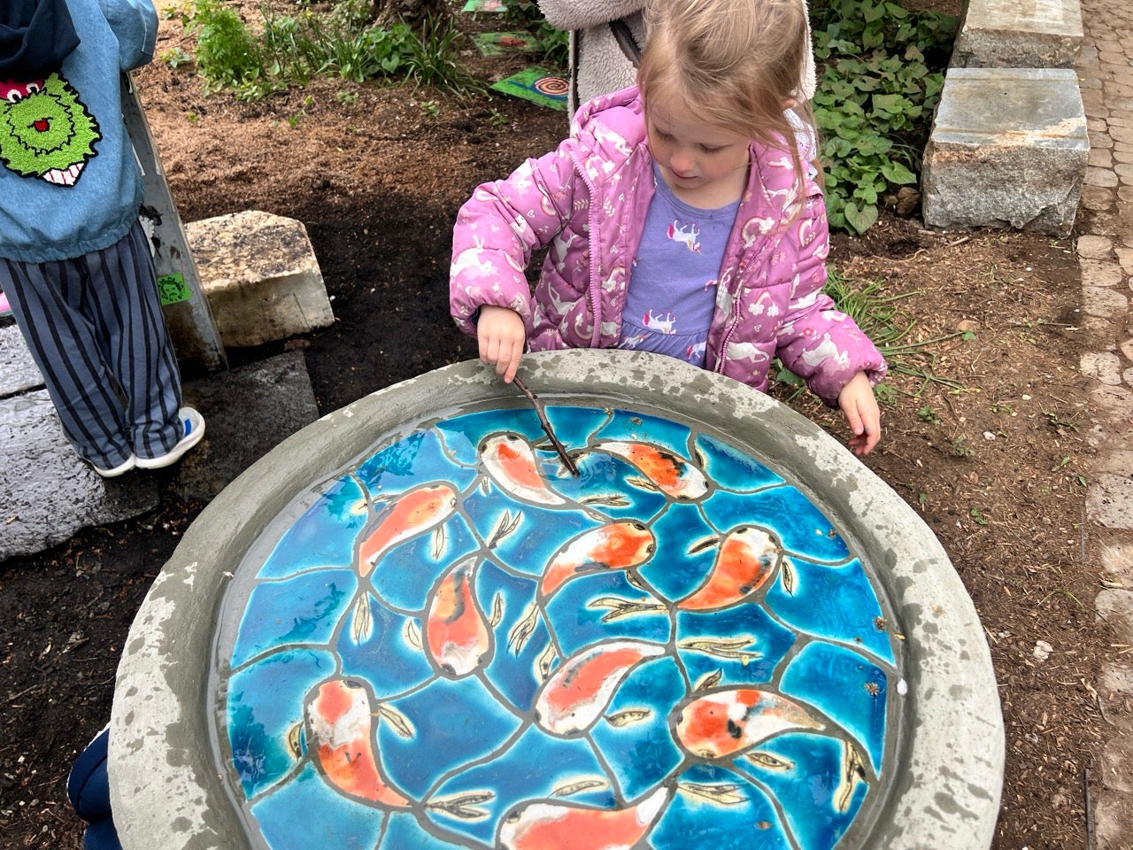 A little girl in a pink unicorn coat sticks her hand in a bird bath painted with colorful koi fish on a bright blue background.