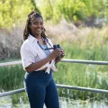 A young Black woman stands outdoors holding a pair of binoculars.