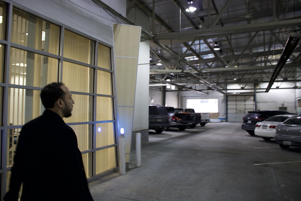 A man walks past glass windows in a well-lit indoor parking garage with several parked cars visible in the background.