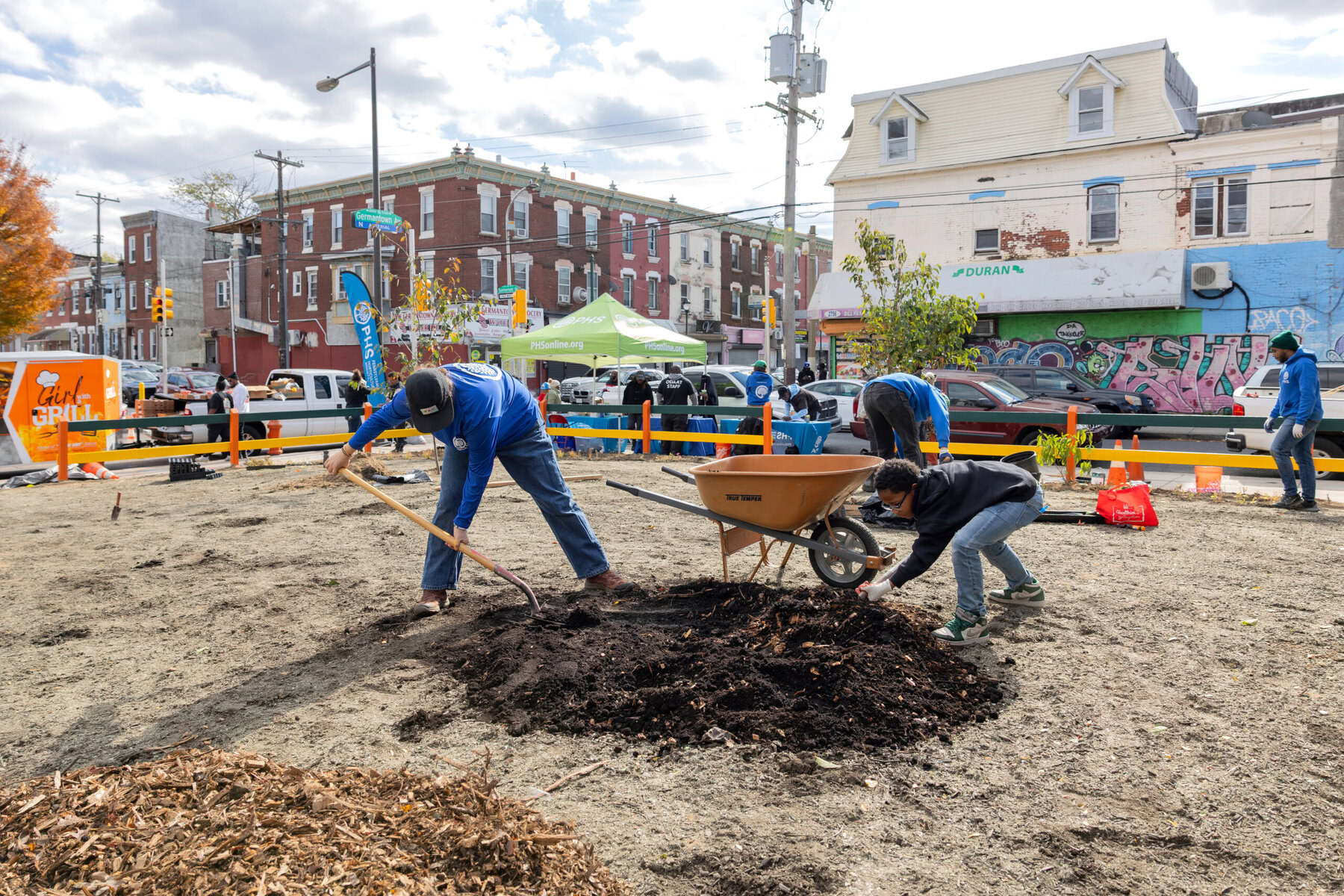 Volunteers and the LandCare team planting a pollinator garden. Two people are placing dirt in an empty lot in front of a wheel barrow with a city block in the background.