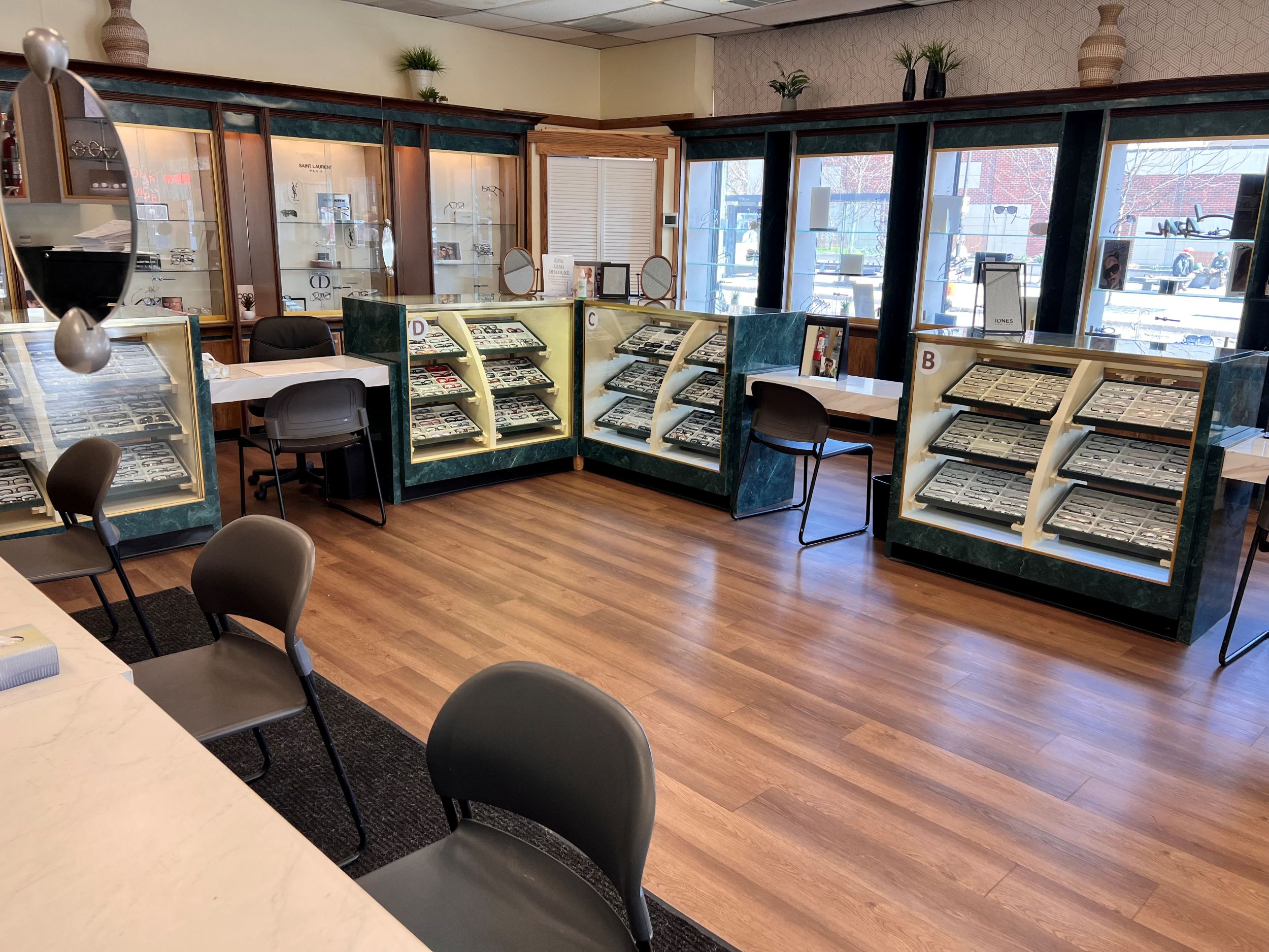 Modern eyewear store interior with display cases of eyeglasses, chairs for customers, wooden flooring, and large front windows showing a street view.