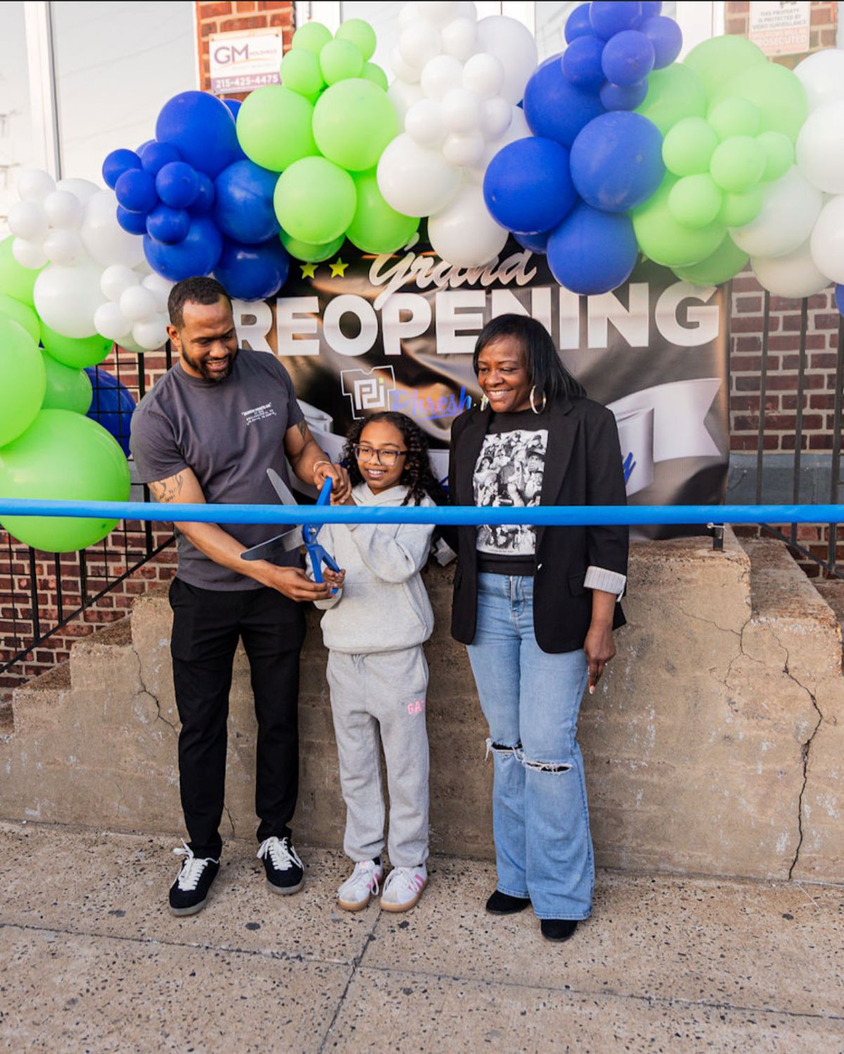 Three people stand in front of a "Grand Reopening" banner with blue and green balloons as a young girl cuts a blue ribbon, while two adults stand on either side of her.