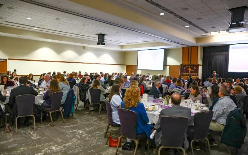 Large conference banquet with attendees seated at round tables facing a speaker and projection screens.
