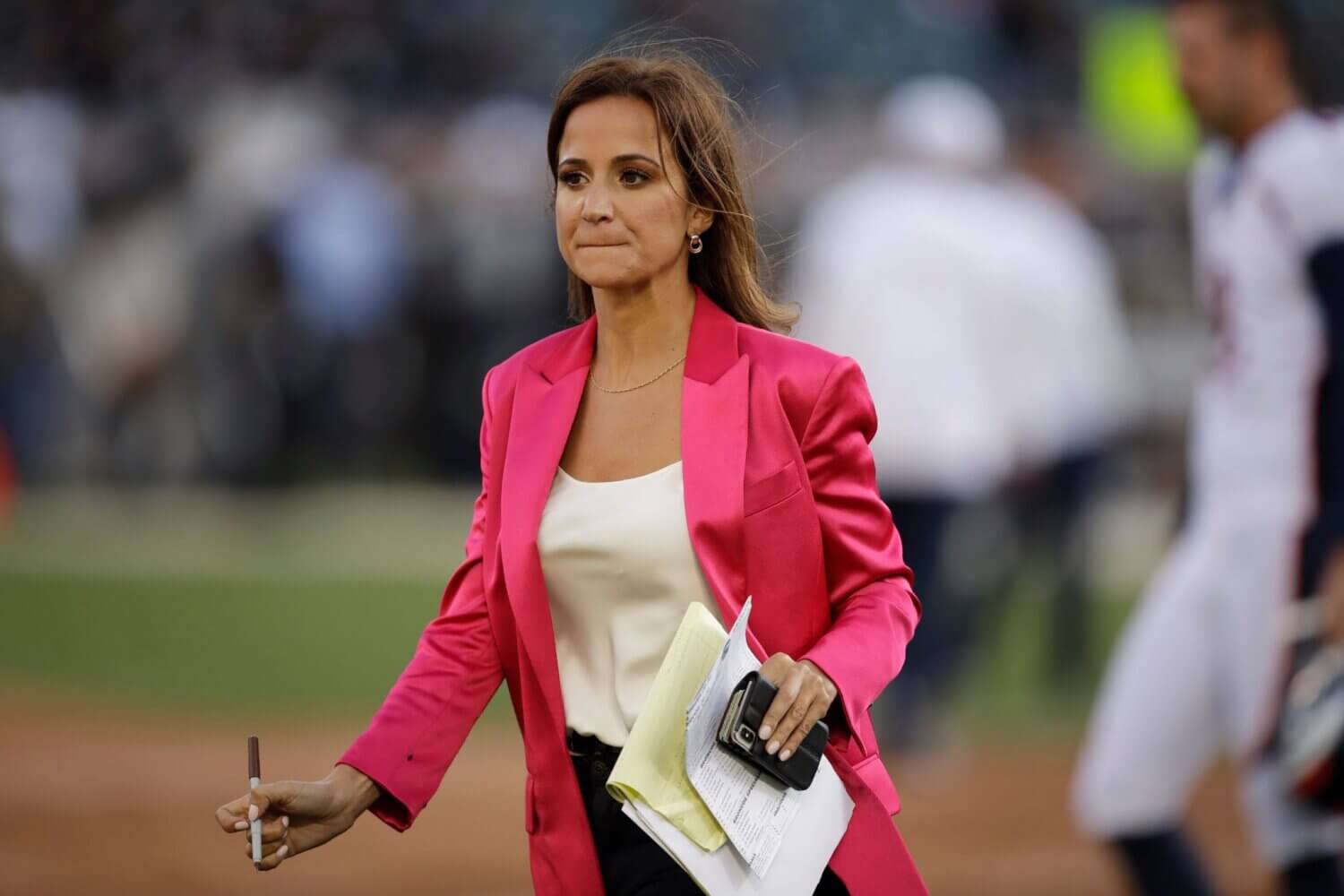 A woman in a pink blazer holding a phone, notebook and pen walks off a sports field