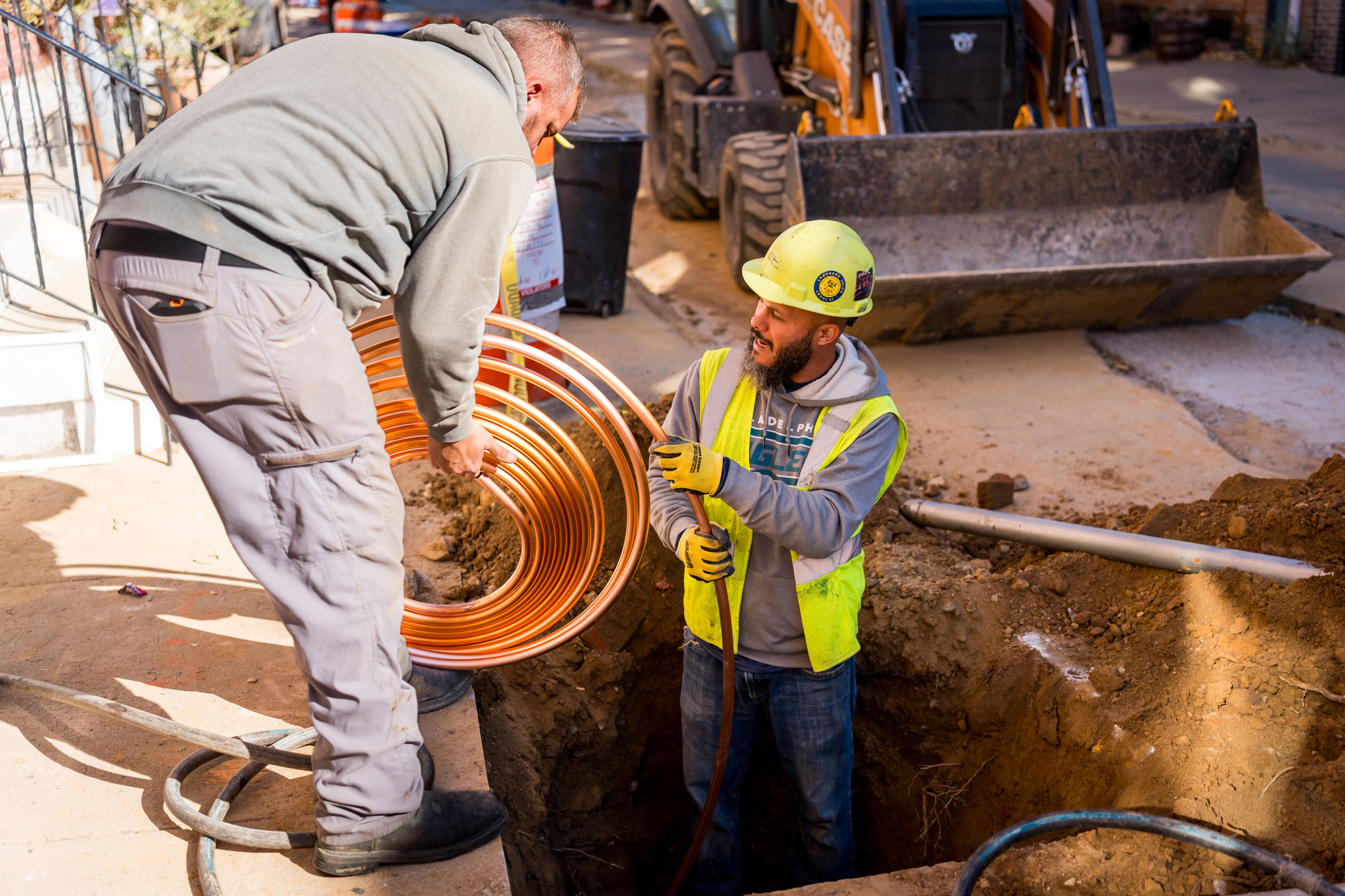 a worker in neon yellow safety gear stands waist-deep in a hole in the sidewalk, guiding copper piping into position from a coil held by another worker outside the hole.