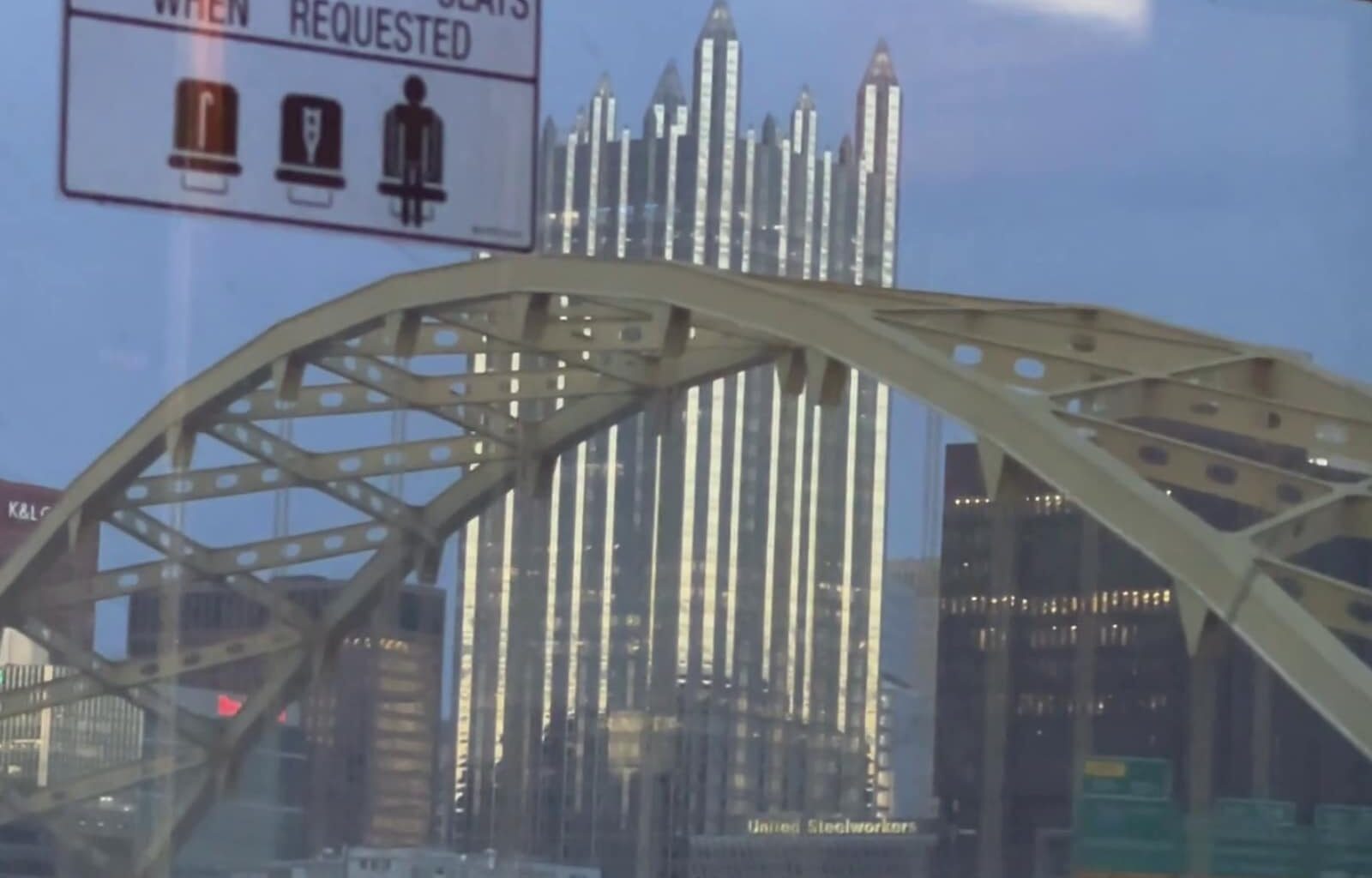Still from the videopoem "Limerent Pittsburgh" shows a public transit view of a yellow arch of the Roberto Clemente Bridge with skyscrapers and signs of Pittsburgh in the distance, including the massive PPG building, which resembles a cathedral.