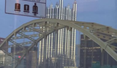 Still from the videopoem "Limerent Pittsburgh" shows a public transit view of a yellow arch of the Roberto Clemente Bridge with skyscrapers and signs of Pittsburgh in the distance, including the massive PPG building, which resembles a cathedral.
