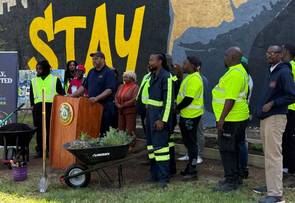 Officials are speaking at a podium surrounded by workers with planting equipment.