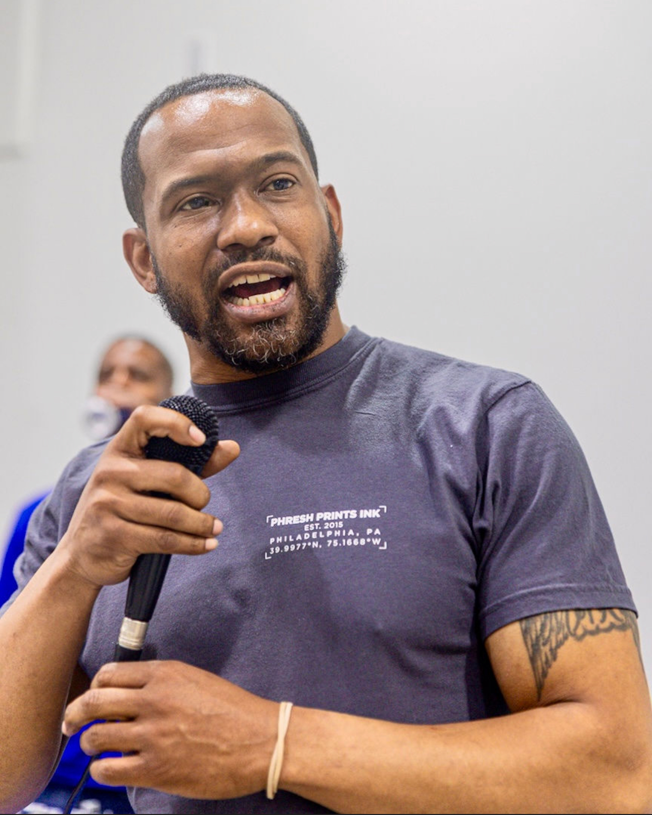 A man with a short beard and mustache speaks into a microphone, wearing a dark t-shirt with text on it. Another person is blurred in the background.