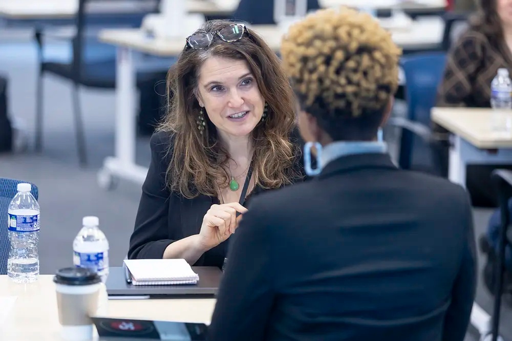 Two women sit at a table in conversation, with notebooks and water bottles in front of them in a classroom or meeting room setting.