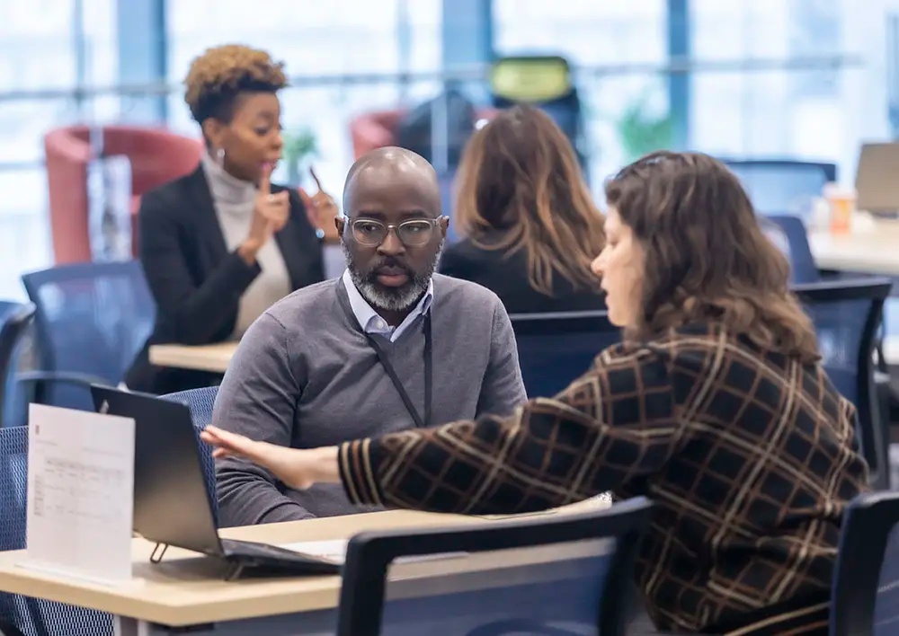 Two people sit at a desk discussing something on a laptop, while others work in the background in a modern office setting.