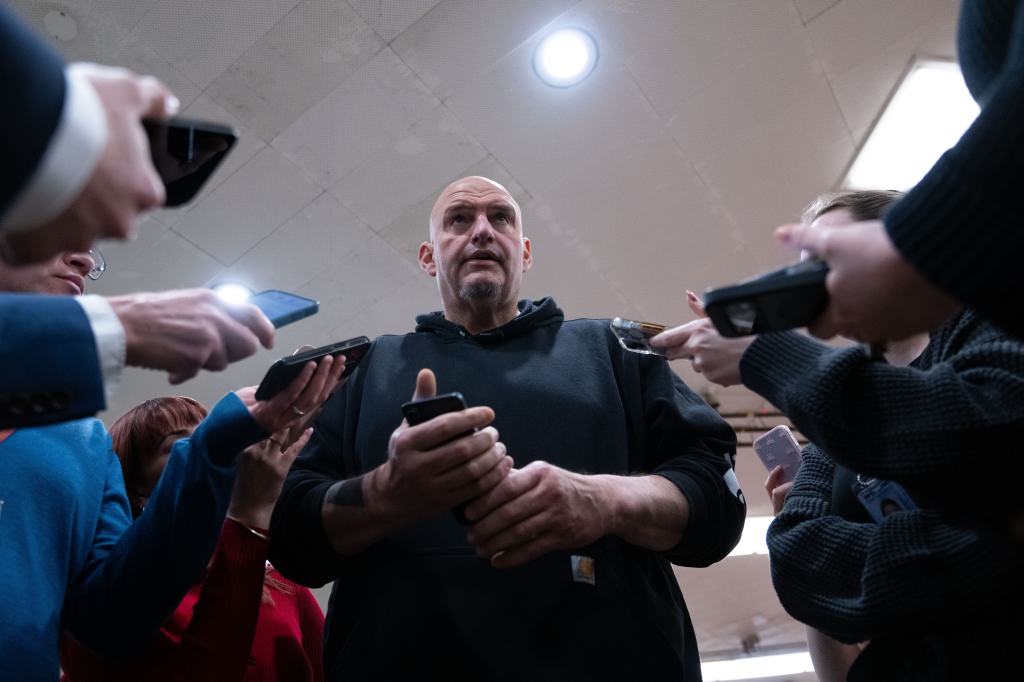Senator John Fetterman, wearing a black hoodie, speaks to reporters who are holding up cell phones to record him.