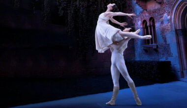 In an Italian-style courtyard, two dancers perform a pas de deux. Zecheng Liang lifts Thays Golz overhead as she tucks one leg into retiré and extends the other back in arabesque. She arches up and back with her arms wide behind her. Both dancers wear white-colored costumes and look up.