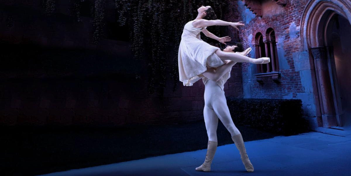 In an Italian-style courtyard, two dancers perform a pas de deux. Zecheng Liang lifts Thays Golz overhead as she tucks one leg into retiré and extends the other back in arabesque. She arches up and back with her arms wide behind her. Both dancers wear white-colored costumes and look up.