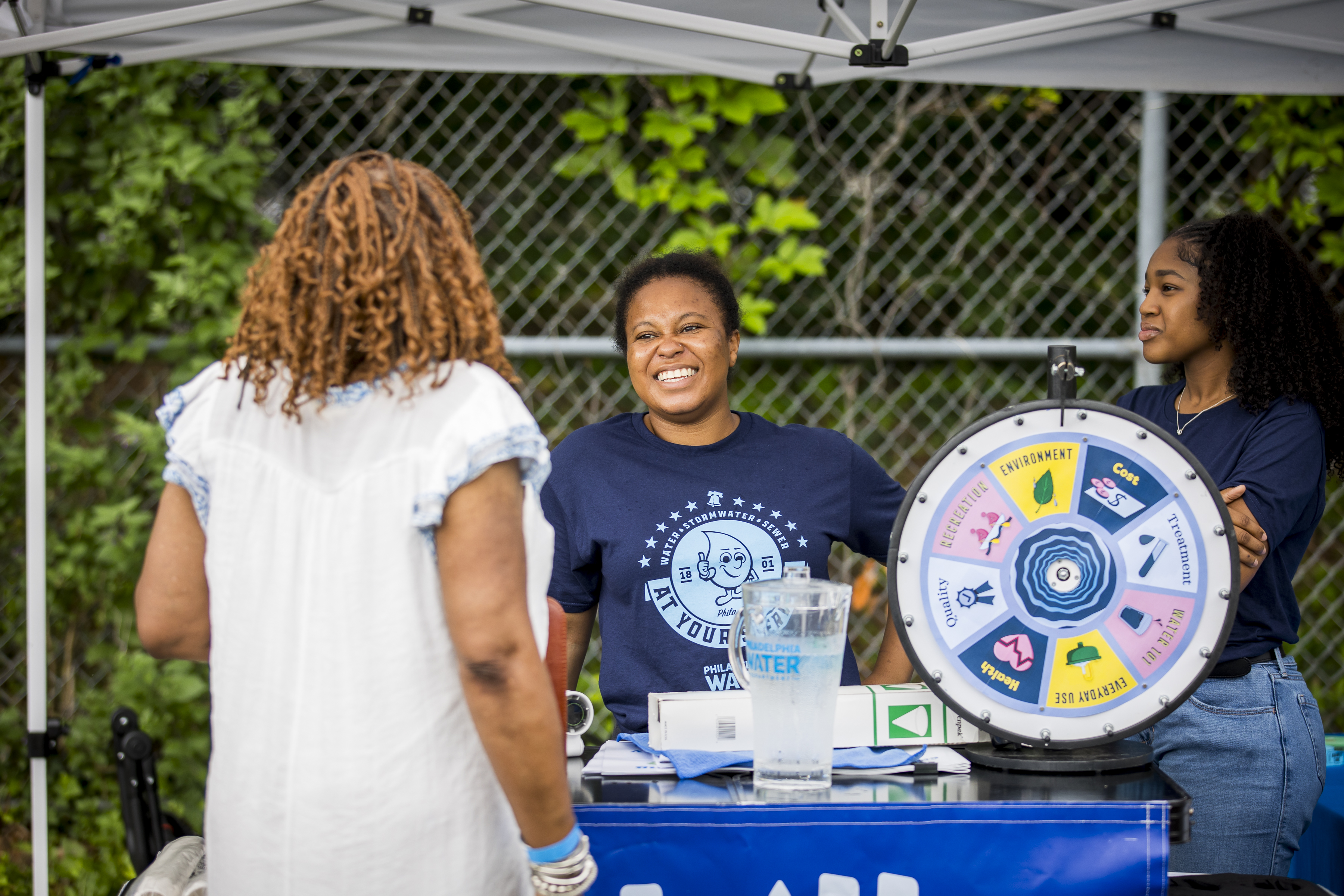 Two smiling PWD employees in dark blue PWD t-shirts stand behind the Philly Water Bar table, talking to a patron with shoulder length curly brown hair and a light blue blouse facing away from the camera. A frosty pitcher of Philly tap water and a "wheel of water" sit on top of the table.