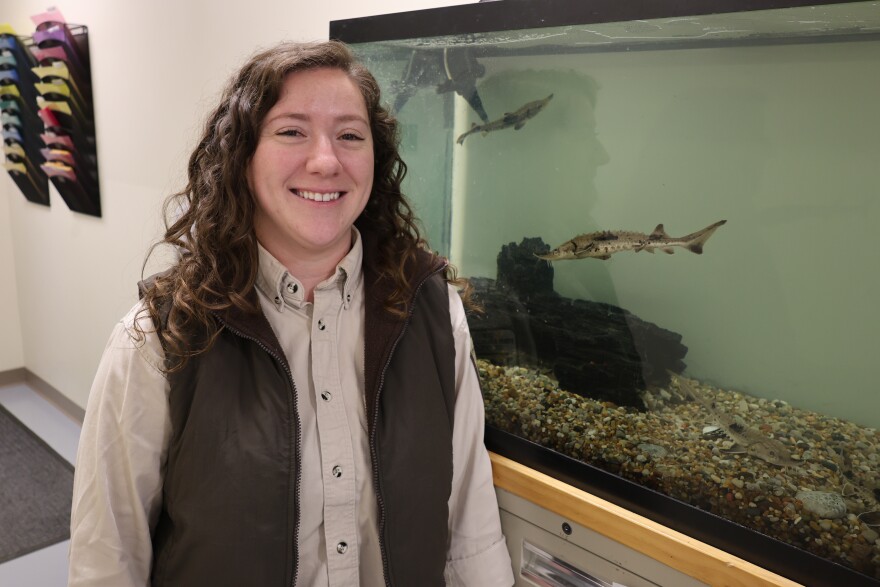 a woman with curly hair stands in front of a fish tank with a baby sturgeon swimming behind her.