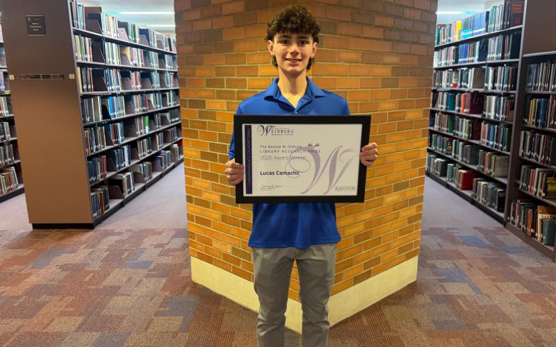 Student standing in a library holding a framed award certificate in front of a brick wall.