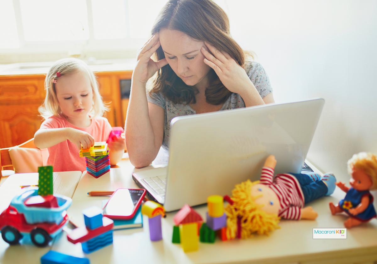 Mom overwhelmed at a laptop while her child plays with toys, showing the mental load of parenting