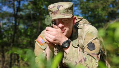 A man in a camouflage military uniform looks through a compass while propped up on his elbows in the grass.
