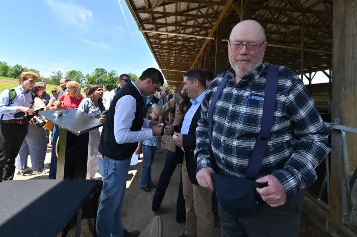 A man in blue flannel shirt and suspenders stands under an overhang at a farm looking at the camera, while the congressman shakes hands in a small crowd behind him.