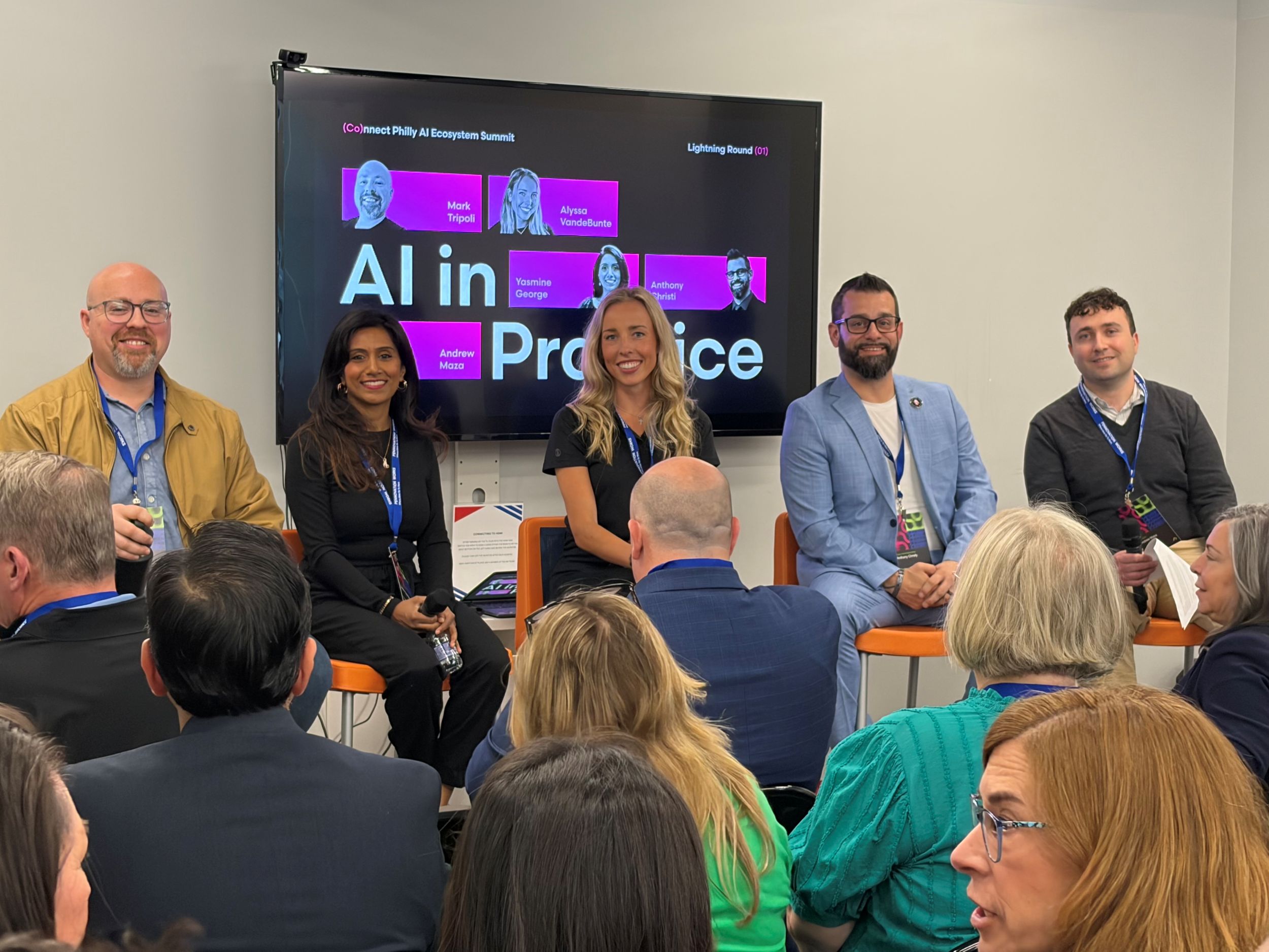 Five panelists sit on stools in front of a screen displaying "AI in Practice," speaking to an audience at a professional event.