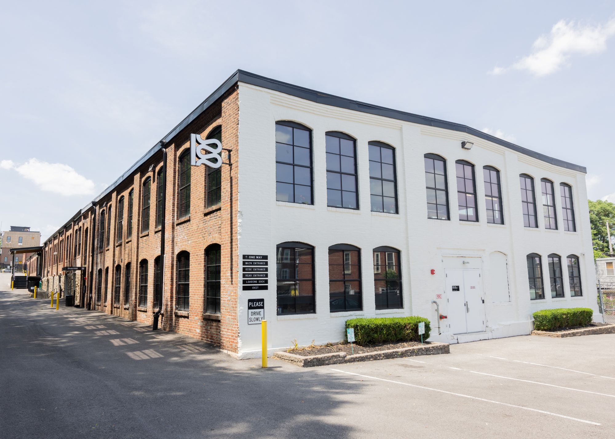 A two-story industrial brick building with large windows, white and red brick facades, and a parking lot in front on a sunny day.