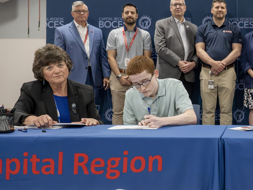 person signs letter while people look on